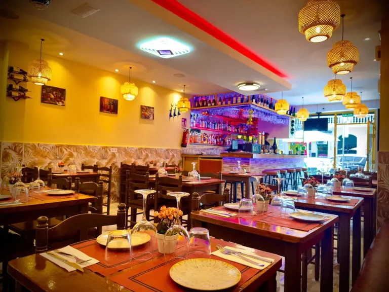 Comedor de Tastari Mediterrani con mesas de madera, lámparas de mimbre y la barra iluminada al fondo