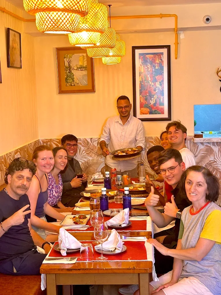 Grupo de clientes sonrientes alrededor de una mesa mientras un camarero sirve una paella
